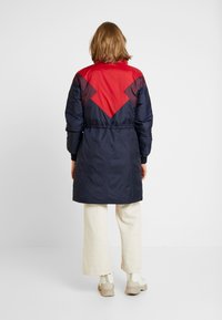 Abrigo acolchado con diseño a bloques de color rojo y navy, cintura ceñida, puños elásticos y una silueta más larga. Tejido suave y brillante.