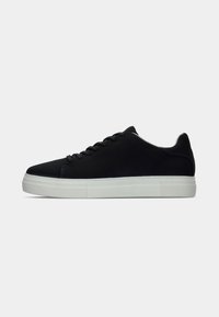 Svart lav sneaker med svarte lisser og tykk hvit gummisåle, vist i sideprofil mot en enkel lys bakgrunn.