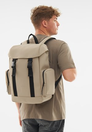 Beige backpack with a structured shape, black accents, front clasps, and side zippered pockets, made from a smooth, durable material.