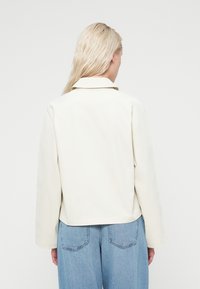 Veste couleur crème en matière lisse, avec un col et une coupe décontractée, associée à un jean denim bleu clair à coupe ample.