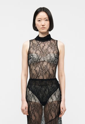 Γυναίκα με κοντά μαύρα μαλλιά που φοράει μαύρο δαντελένιο φόρεμα χωρίς μανίκια πάνω από γκρι τοπ bandeau και εσώρουχα ψηλόμεσα μπροστά σε ένα απλό φόντο.