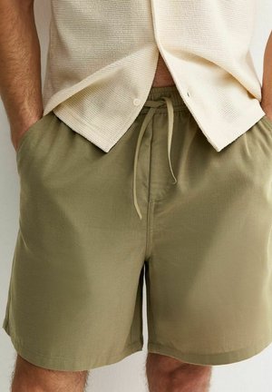 Man met licht beige, gestructureerd shirt met korte mouwen en olijfgroene shorts met trekkoord, met handen in de zakken.
