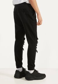 Czarne dżinsy o slim fit, z postrzępionymi detalami po bokach, elastycznymi mankietami i dwiema tylnymi kieszeniami, połączone z czarnymi trampkami i białymi skarpetkami.