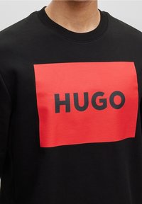 Sudadera negra con un gran gráfico rectangular rojo que muestra la palabra "HUGO" en una fuente negra y audaz. Cuello acanalado y textura suave.