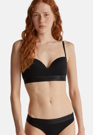 Vrouw met lang rood haar, gekleed in een zwarte beha en bijpassende onderbroek met "United Colors of Benetton"-merk op de banden.