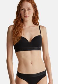 Mladá žena s dlouhými rudými vlasy, oblečená v černé podprsence a odpovídajícím černém prádle s logem "United Colors of Benetton".