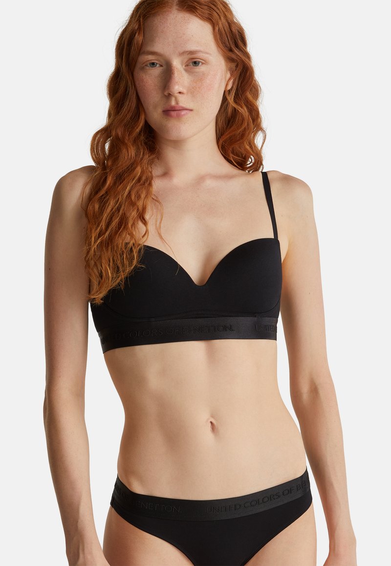 Mladá žena s dlouhými rudými vlasy, oblečená v černé podprsence a odpovídajícím černém prádle s logem "United Colors of Benetton".