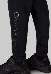 Pantaloni sportivi neri con logo Calvin Klein in argento riflettente, caratterizzati da un design affusolato e bordi a costine. Abbinati a scarpe da ginnastica nere.