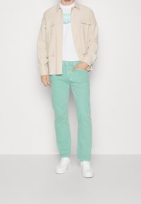 Ljus beige knäppskjorta över en vit t-shirt, matchad med mintgröna jeans och vita sneakers. Slim-fit design med fickor.