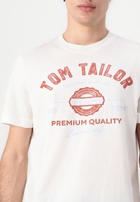Hvit bomull T-skjorte med en teksturert rød trykk som har teksten "TOM TAILOR" og "PREMIUM QUALITY", komplementert av et sirkulært merke-design.