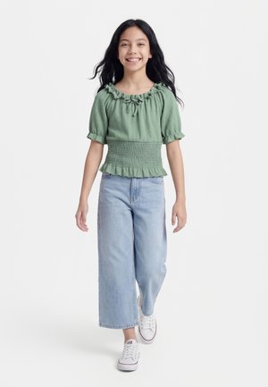 Uśmiechnięta dziewczyna z długimi czarnymi włosami, idąca, ubrana w zieloną bluzkę z falbanami, jasnoniebieskie szerokie spodnie jeansowe i białe trampki.