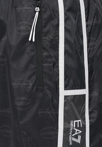 Chaqueta ligera negra con rayas blancas en los lados, bolsillo con cremallera y un patrón sutil de logotipos. Presenta cordón ajustable y textura suave.