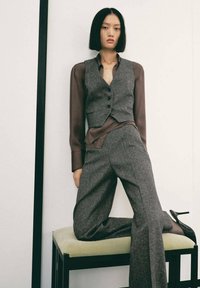 Grauer Wollweste mit Knöpfen, kombiniert mit einer braunen, seidenen Bluse und grauen Hosen. Das Model sitzt auf einem grünen gepolsterten Hocker. Schwarze Absatzschuhe.