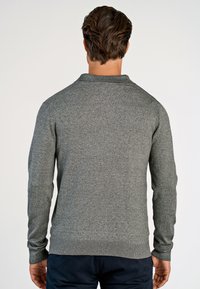 Mand med brunt hår, vendt bort fra kameraet, iført en langærmet grå trøje og mørke bukser, mod en ensfarvet lys baggrund.