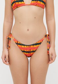 Bikini-Höschen mit Seitenschnürung, das ein Batikmuster in Orange, Gelb und Schwarz aufweist. Hergestellt aus dehnbarem Stoff mit kontrastierenden Nähten und Perlen.