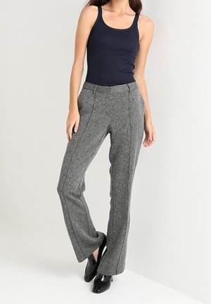 Kobieta ubrana w czarny, bezrękawnik, szare, dopasowane, szerokie spodnie oraz czarne, błyszczące buty na obcasie, stojąca z jedną nogą założoną na drugą.