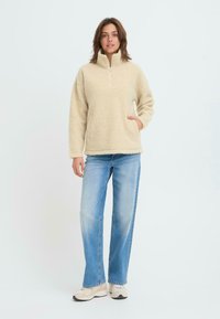 Beżowy polarowy sweter z półzbiegowym kołnierzem i bocznymi kieszeniami, zestawiony z jasnoniebieskimi dżinsami prostymi oraz obuwiem sportowym w neutralnych odcieniach.
