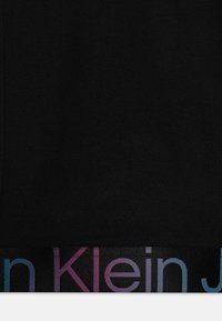 Musta kangas sileällä pinnalla, jossa on värikäs, häilyvä logonauha helman kohdalla, jossa lukee "Calvin Klein."