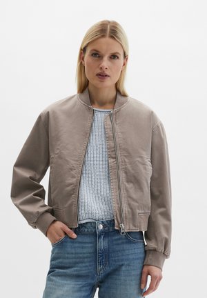 Blonde Frau trägt eine taupefarbene Bomberjacke über einem hellblauen Strickpullover, mit der Hand in der Tasche, stehend vor weißem Hintergrund.