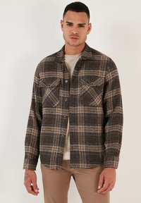 Tamsių tonų checkered marškiniai su smėlio spalvos akcentais, turi užsegimą mygtukais, dvi krūtinės kišenes ir laisvą siluetą. Pagaminti iš tekstūruoto audinio.