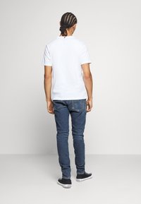 Bílé tričko s krátkým rukávem zobrazené zezadu, doplněné tmavě modrými džíny. Džíny mají zadní kapsy a úzký střih.