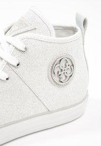 Biała niska tenisówka z płaskimi białymi sznurowadłami, teksturowaną cholewką oraz okrągłą srebrną naszywką z logo po boku.