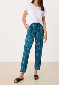 Teal gekleurde bloemenpatroon broek met een elastische tailleband en zijzakken, gecombineerd met een losse witte t-shirt en zwarte slippertjes.