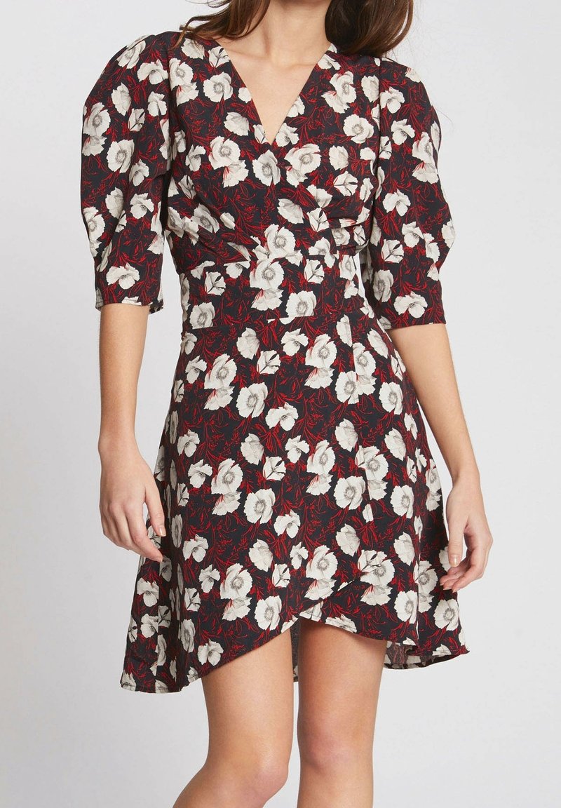 Robe portefeuille noire avec imprimé floral blanc, accents feuilles rouges, encolure en V, manches trois-quarts bouffantes et ourlet asymétrique sur une femme.