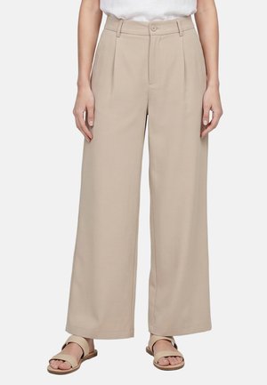 Beige Hosen mit weitem Bein und vorderen Falten, Gürtelschlaufen, Knopf- und Reißverschluss, getragen mit beigen flachen Sandalen an einer stehenden Person.
