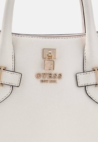 Biała torebka ze skóry ziarnistej z złotymi okuciami, wyposażona w zapięcie na zatrzask oraz wytłoczone logo "GUESS EST 1981" na przedniej stronie.