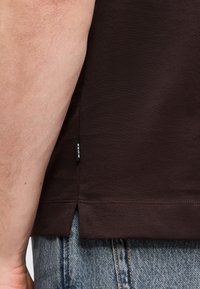 Πλάγια όψη ενός ατόμου που φοράει σκουρόκαφέ πουκάμισο με μια μικρή ετικέτα "BOSS" κοντά στο τελείωμα και ανοιχτόχρωμο τζιν παντελόνι.