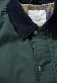 Chaqueta de tela verde con un cuello de pana azul marino, cierre a presión y forro de cuadros. La etiqueta dice "Seasalt Cornwall."