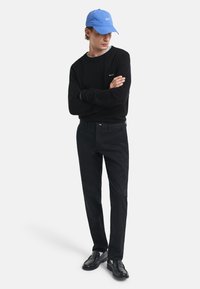 Suéter negro de cuello redondo, pantalones negros, mocasines negros y una gorra azul con texto blanco. El modelo está de pie con los brazos cruzados frente a un fondo blanco.