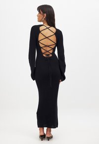 Robe en tricot noir avec des manches longues, un décolleté profond dans le dos avec un laçage croisé, et une silhouette cintrée qui tombe jusqu'aux chevilles.