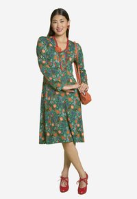 Grünes Blumenkleid mit langen Ärmeln, das mit orangefarbenen und weißen Blumen verziert ist, kombiniert mit roten Akzenten und einer kleinen orangefarbenen Handtasche. Rote Schuhe vervollständigen den Look.