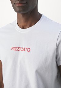 Hvid bomulds t-shirt med rund hals og korte ærmer, med "PIZZICATO" trykket i fede røde bogstaver på brystet.