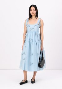 Vestido sem mangas azul claro com decote quadrado, camadas com babados e laços de tecido. Usado com sapatos rasos pretos com bolinhas e uma bolsa com tachas.