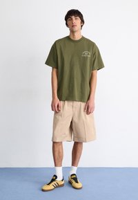 Camiseta de algodón verde oliva con mangas cortas y escote redondo, combinada con pantalones cortos de algodón beige hasta la rodilla y zapatillas amarillas y negras.