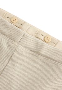 Beżowy materiał prążkowany z delikatnym połyskiem, z dwoma brązowymi guzikami w talii i przeszytymi akcentami wzdłuż pasa.