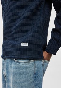 Tengerészkék texturált ing méhsejt mintával, fekete gombokkal és címkével, világoskék klasszikus szabású farmernadrággal párosítva.