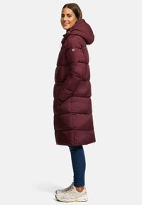 Lang bordeauxkleurige pufferjas met een capuchon, voorzien van horizontale stiksels en zijzakken. Gepaard met blauwe jeans en lichte sneakers.