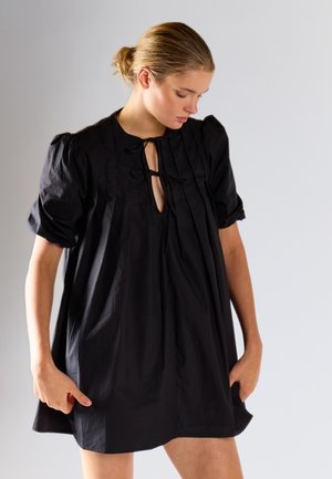 Giovane donna con capelli biondi raccolti in uno chignon, indossa un abito nero largo con maniche a sbuffo e dettagli annodati, guarda verso il basso di lato.