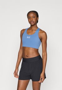 Μπλε αθλητικό crop top με στρογγυλή λαιμόκοψη και λογότυπο, σε συνδυασμό με μαύρο σορτς με ελαστική μέση, άνετη γραμμή και πιέτες.