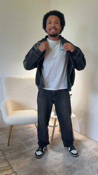 Czarna kurtka z błyszczącymi akcentami, biała koszulka, ciemne dżinsy i czarne trampki z białymi detalami. Umieszczony w przytulnym wnętrzu.