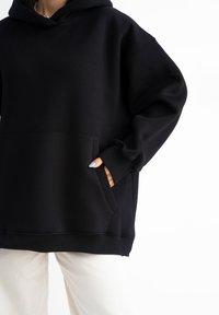 Felpa nera oversize realizzata in morbido misto cotone. Caratterizzata da una tasca frontale, polsini a coste e un grande cappuccio. Indossata con pantaloni di colore chiaro.