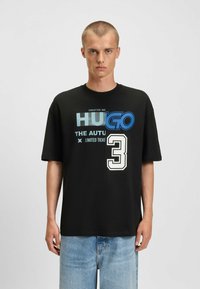 Camiseta de algodón negra con texto gráfico y el número '3' en azul y blanco. Manga corta, corte relajado y diseño de cuello redondo.