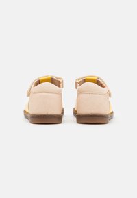 Un paio di sandali per bambini beige e gialli con cinghie alla caviglia, visti da dietro su uno sfondo bianco.