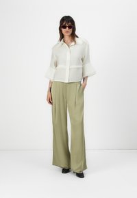 Chemise blanche cropped avec des accents à franges et un devant à boutons, associée à un pantalon large olive et des mules noires.
