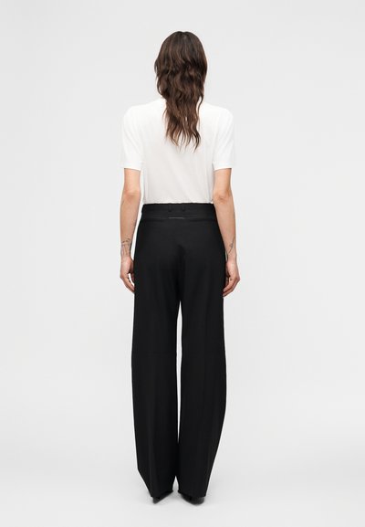 Γυναίκα με μακριά καστανά μαλλιά φοράει λευκό τοπ με κοντά μανίκια και μαύρο ψηλόμεσο παντελόνι με φαρδιά μπατζάκια, στέκεται με την πλάτη προς τα εμάς σε απλό φόντο.