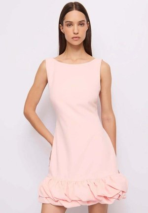 Giovane donna con capelli castani lisci che indossa un vestito rosa pallido senza maniche con orlo arricciato, in piedi contro uno sfondo chiaro e uniforme.